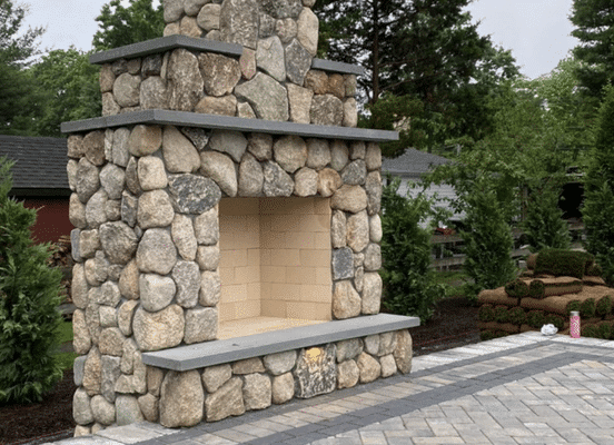 Large outdoor stone fireplace crafted with round fieldstones and a dark stone hearth, set in a backyard patio.