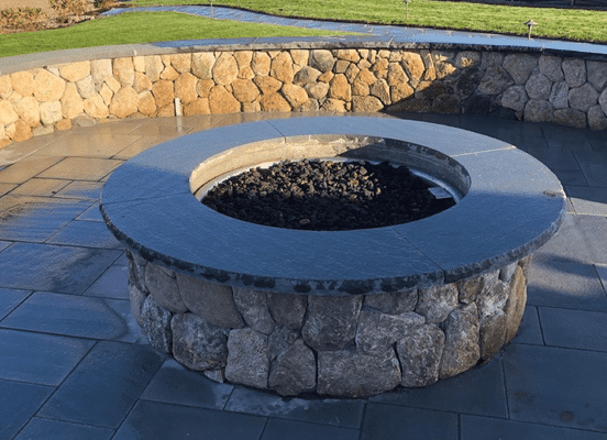 Round outdoor fire pit made of stacked natural fieldstone with a dark top cap, built on a stone patio.